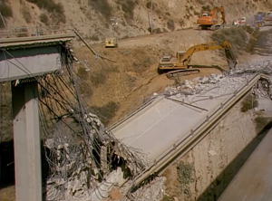 A partially collapsed highway bridge lies in ruins, with twisted rebar and broken concrete visible. Construction equipment, including excavators and bulldozers, are positioned nearby on a dirt road as part of a demolition or reconstruction effort. The scene is set in a hilly, dry landscape.